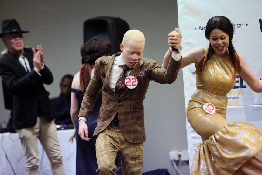 Two albinism pageant contestants hold hands and dance in the middle of a stage as others watch on and clap.