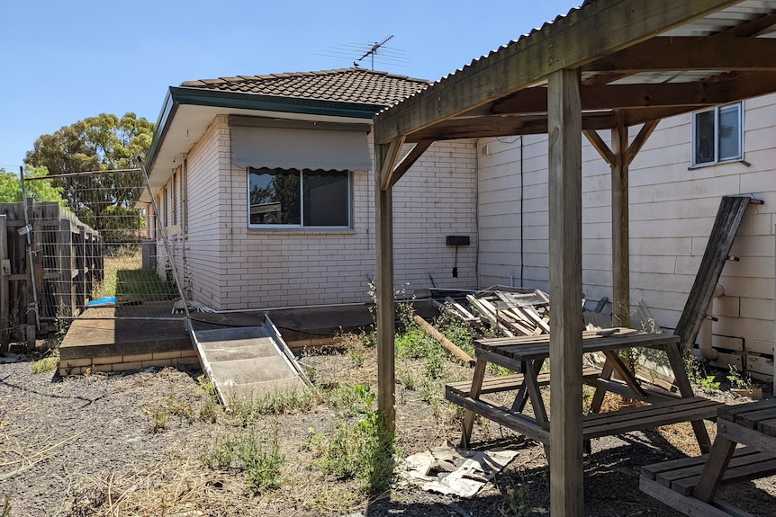 A backyard with overgrown and dead grass, piles of timber, broken metal fencing, and a ramp.