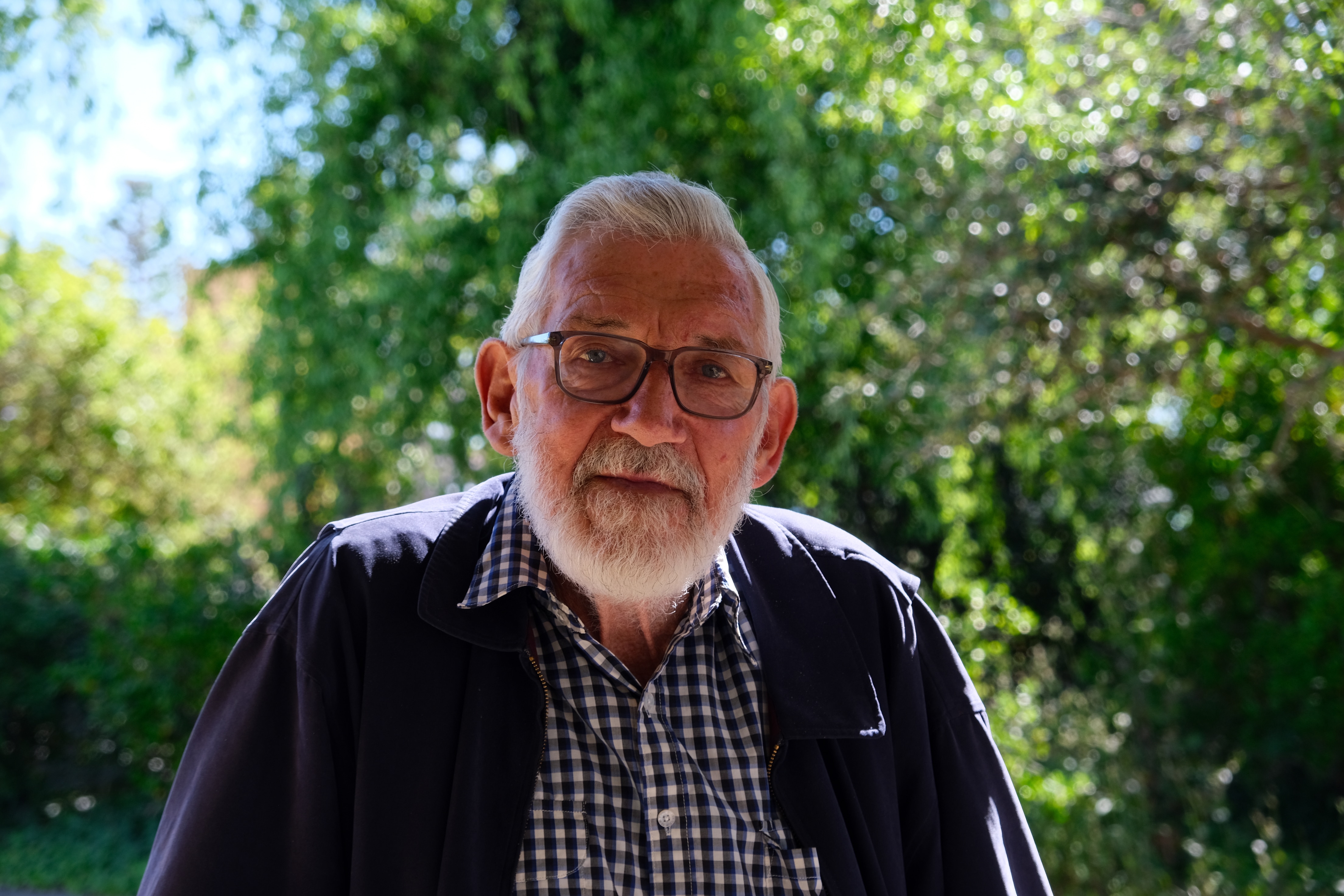 A man wearing a checked shirt and blue jacket looks straight ahead. He has a grey beard and is wearing glasses