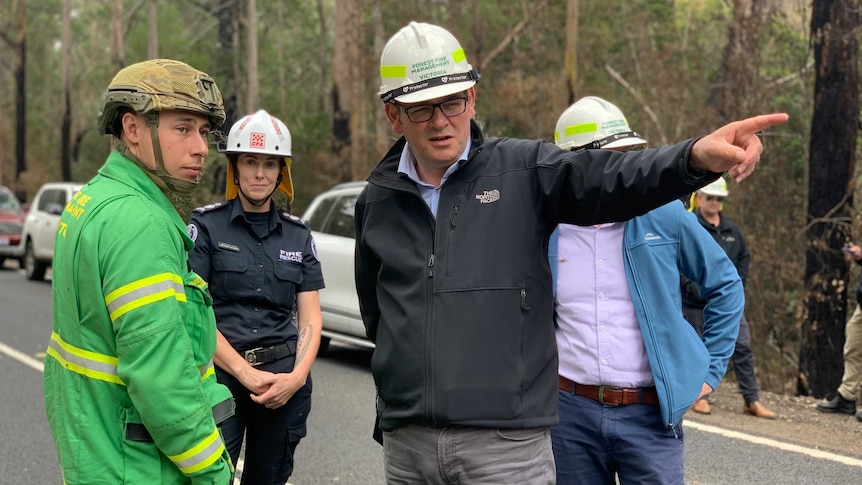 Daniel Andrews wearing a white hard hat points into the distance while speaking to an emergency official.