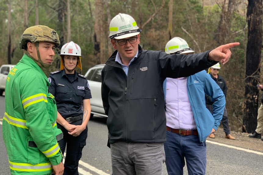 Daniel Andrews wearing a white hard hat points into the distance while speaking to an emergency official.