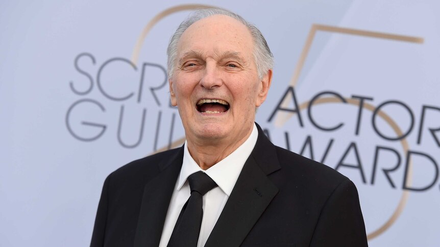 Alan Alda grins against SAG awards red carpet backdrop. He wears a black suit and tie and white shirt.