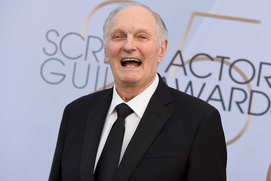 Alan Alda grins against SAG awards red carpet backdrop. He wears a black suit and tie and white shirt.