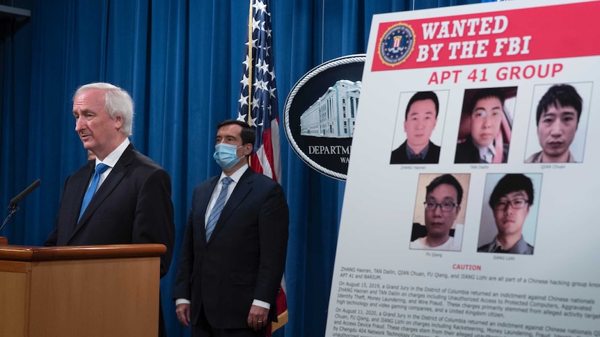 Deputy Attorney General Jeffrey Rosen speaks from a podium next to a poster with five faces of the Chinese hackers.