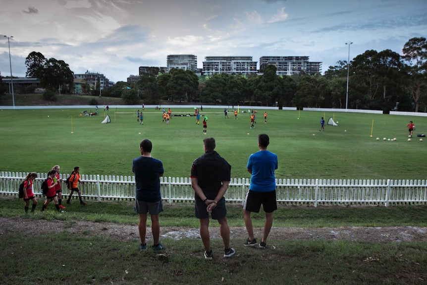 Three men stand near an oval, as apartment buildings can be seen in the distance