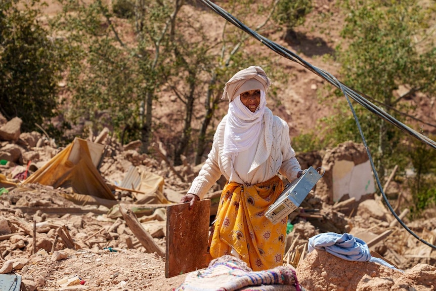a woman surrounded by rubble, carrying two pieces of wood.