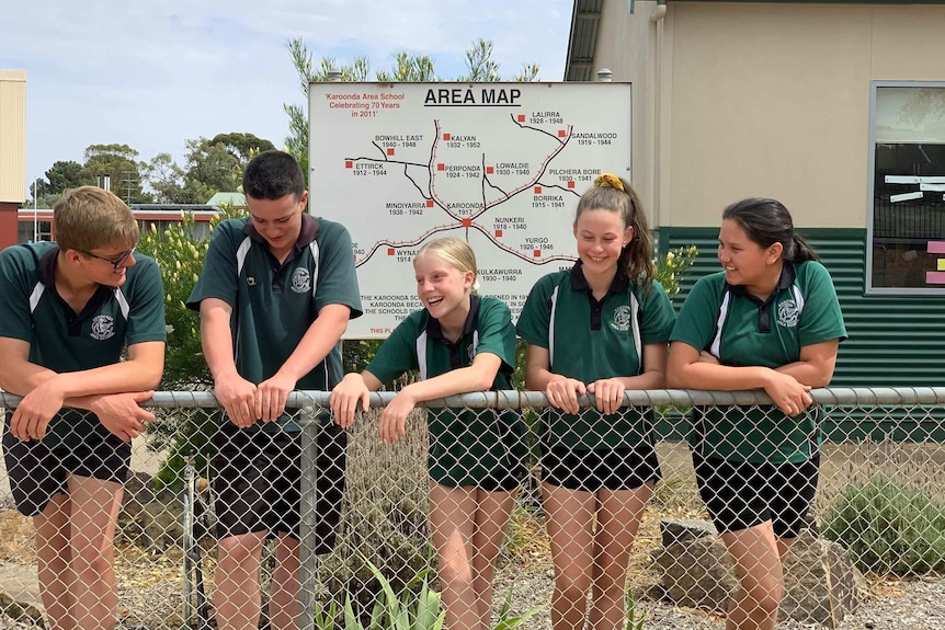 Five students stand in front of a map, leaning on a wire fence. They are smiling.