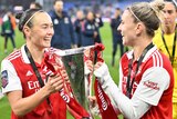 Two soccer players wearing red and white hold a silver trophy while smiling at each other