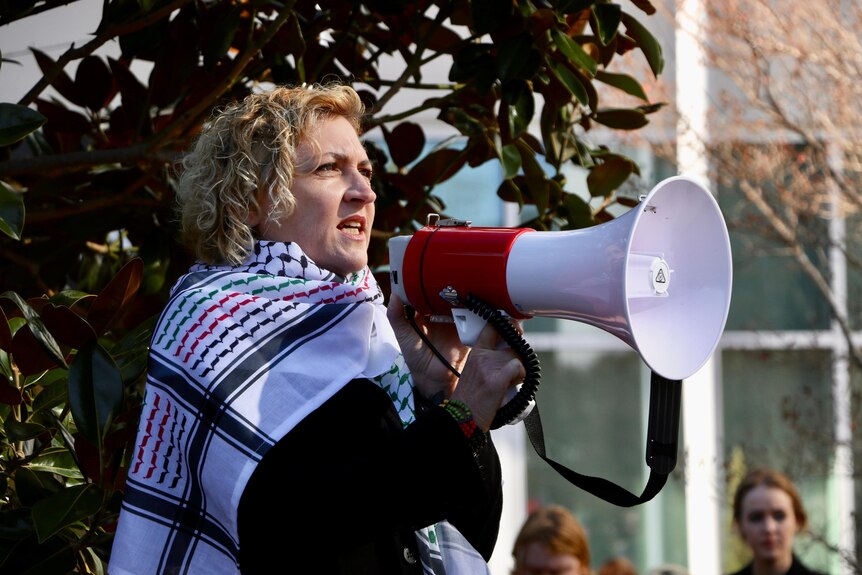 ACT Greens minister Emma Davidson, wearing a  keffiyeh, speaks into a megaphone. 