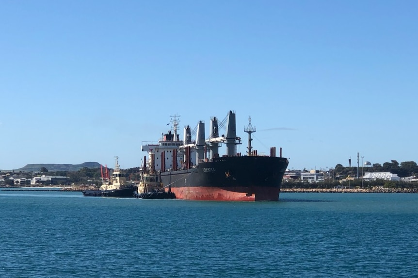 Two tug boats guide a cargo ship, the Liberty C, into the Geraldton port on a sunny, cloudless day.