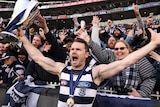 Patrick Dangerfield holds his arms out while holding the premiership cup and yells, standing in front of Cats fans