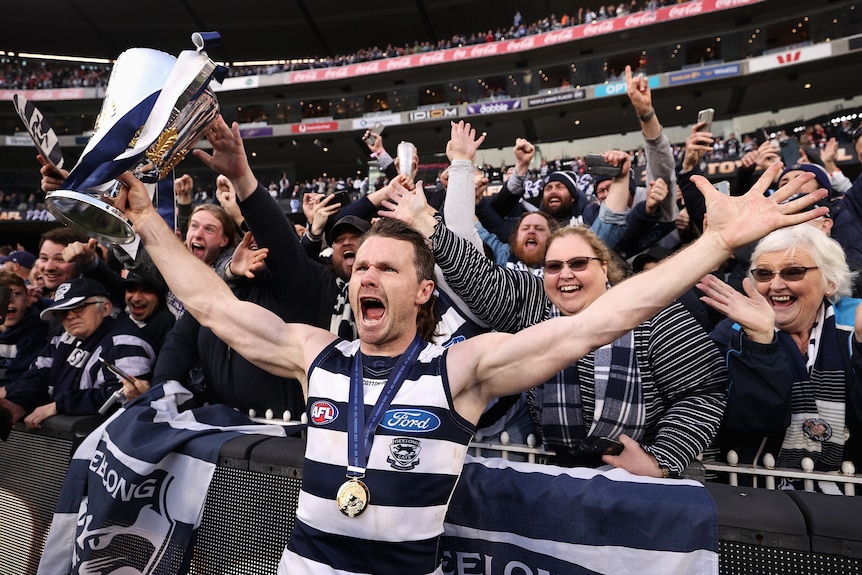 Patrick Dangerfield holds his arms out while holding the premiership cup and yells, standing in front of Cats fans