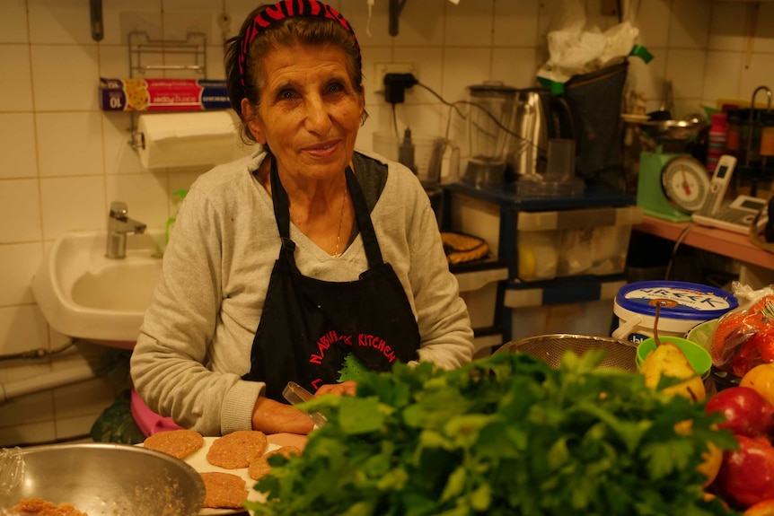 An elderly Lebanese woman standing in the kitchen of a cafe, with fruit and celery in front of her.