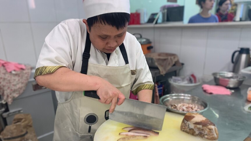 Chef holds large chopping knife, cutting roast pork on chopping board in a restaurant while customers wait