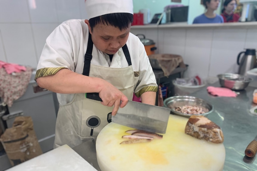 Chef holds large chopping knife, cutting roast pork on chopping board in a restaurant while customers wait