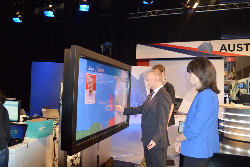 Antony Green in TV studio touching touch screen with other production crew watching on.