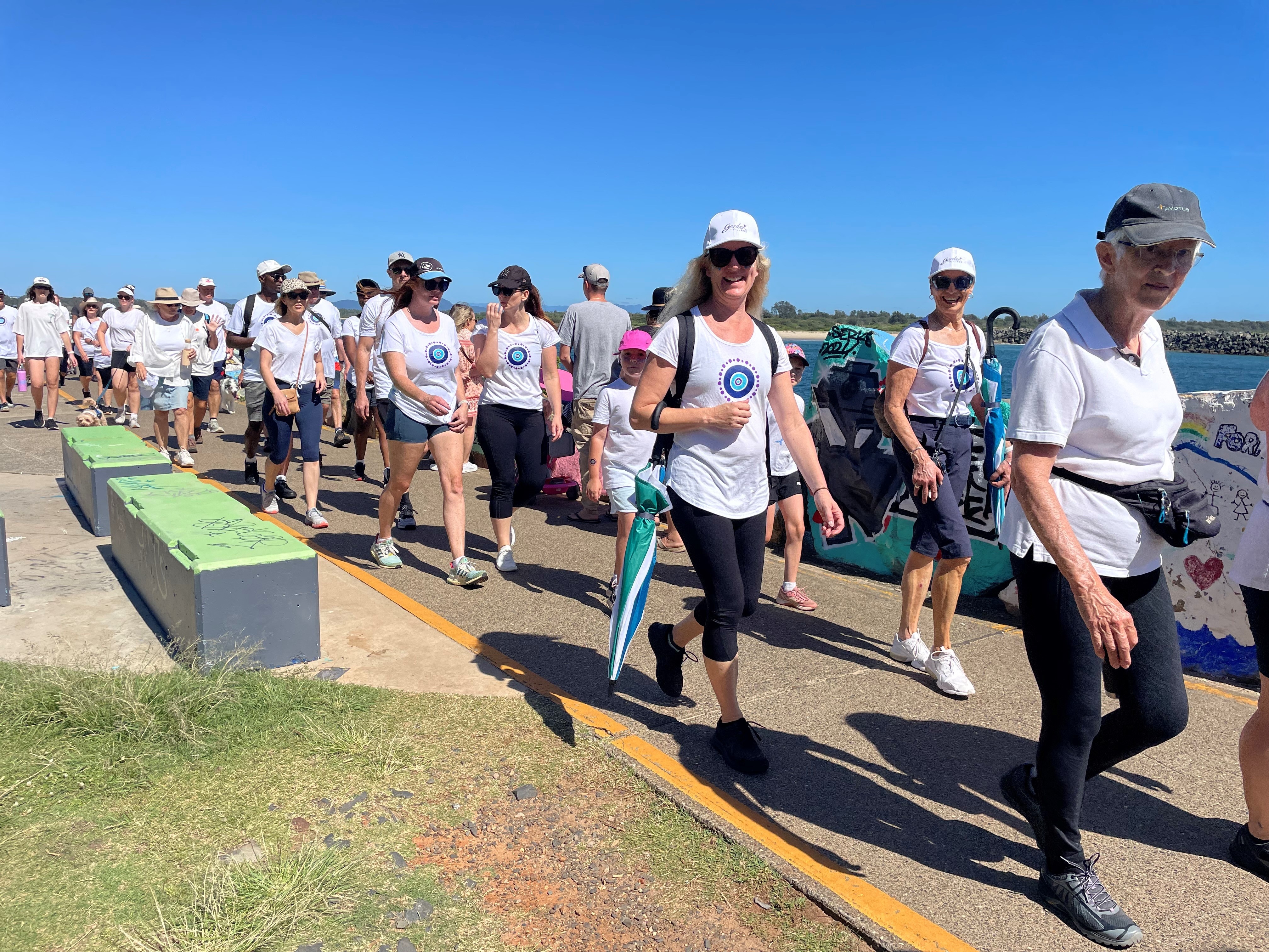 A line of people in white t-shirts walk along a path next to a river, under a blue sky.