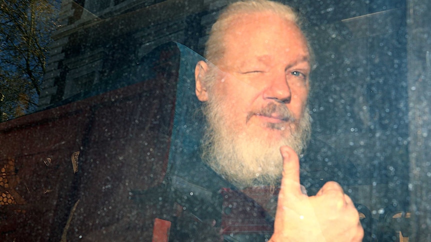 WikiLeaks founder Julian Assange is seen through a dirty police van window as he winks and throws a thumb in the air.