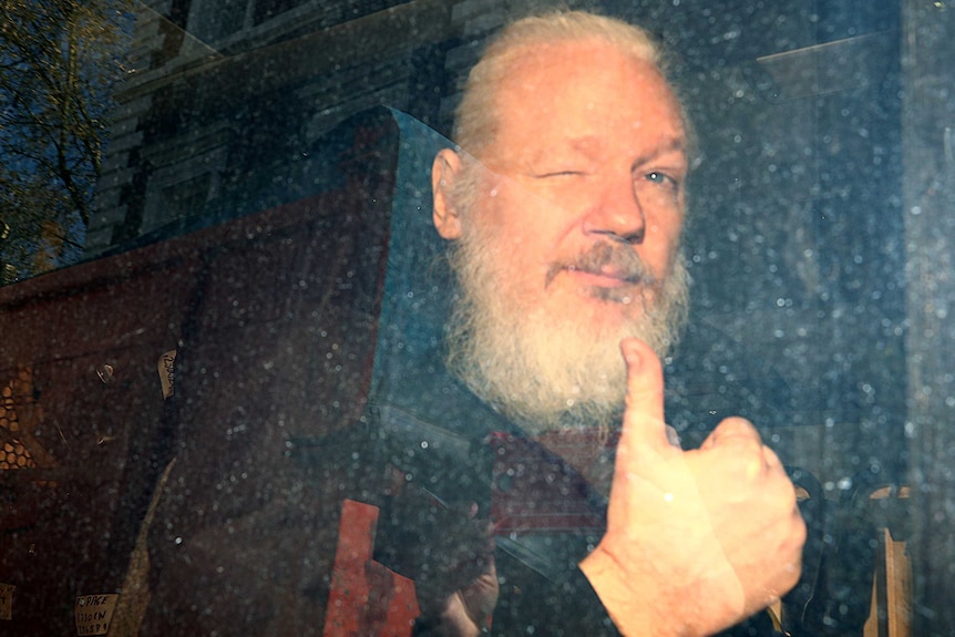 WikiLeaks founder Julian Assange is seen through a dirty police van window as he winks and throws a thumb in the air.