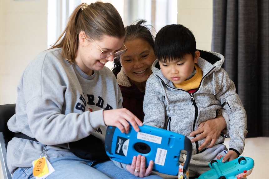 Three people sit smiling looking at an AAC device.