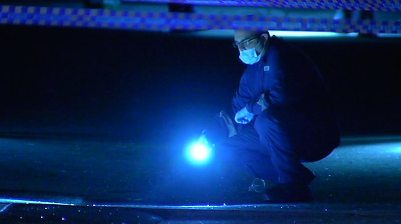 A forensic police officer wearing a face mask shines a torch at a crime scene marked off with police tape.