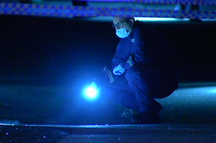 A forensic police officer wearing a face mask shines a torch at a crime scene marked off with police tape.