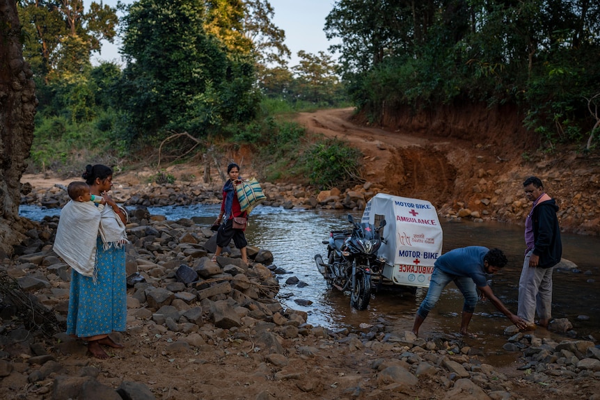 A woman holding a baby watches as two men move rocks so a motorbike ambulance can pass through a river. 