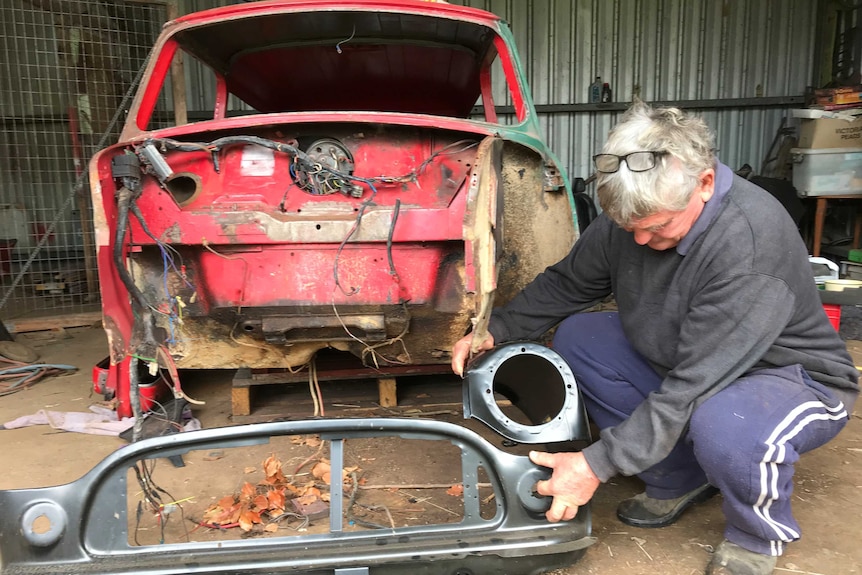 An elderly man crouches down in a farm shed as he works on restoring an old car