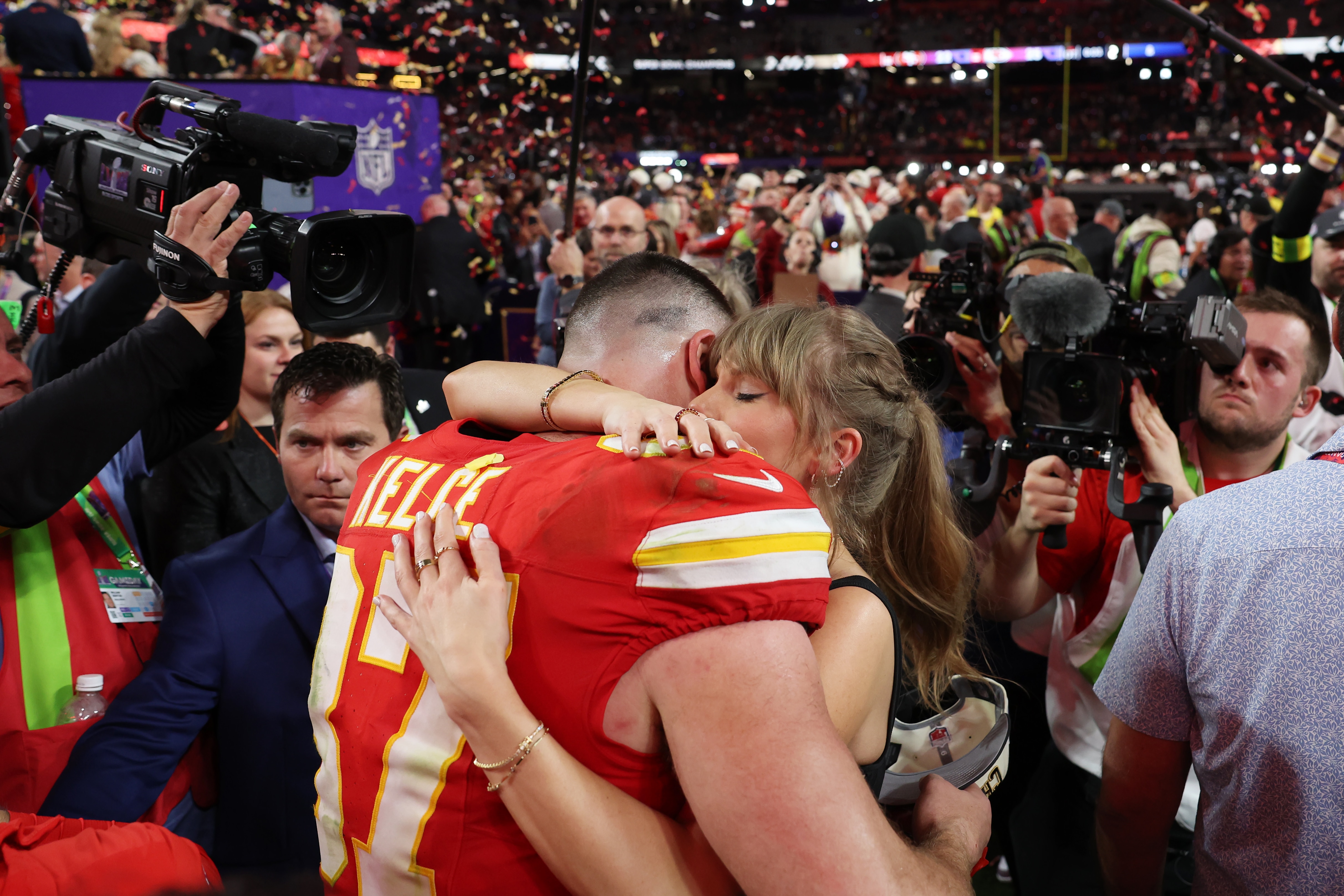 A male NFL player engages hugs girlfriend inside a stadium, with cheering fans, cameras and confetti surrounding them
