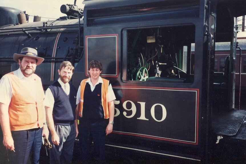 Three men in railway worker uniforms stand in front of a steam train.