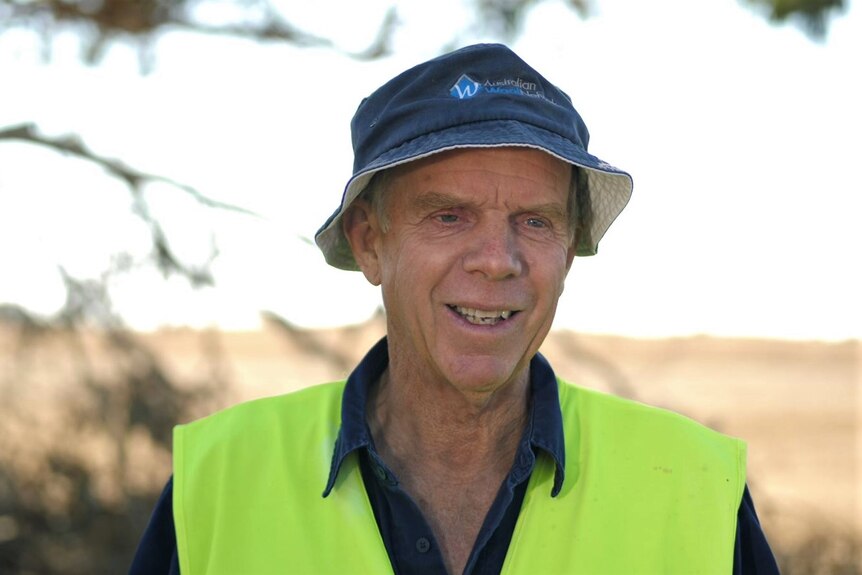 An elderly man wearing a navy blue bucket hat and bright high vis vest smiles. 