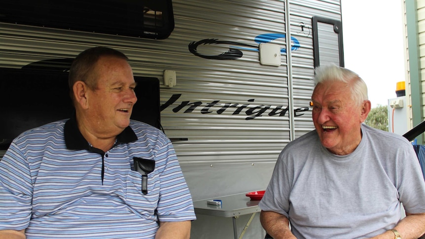 Two men sitting in camping chairs laugh, looking at each other, out the front of a caravan