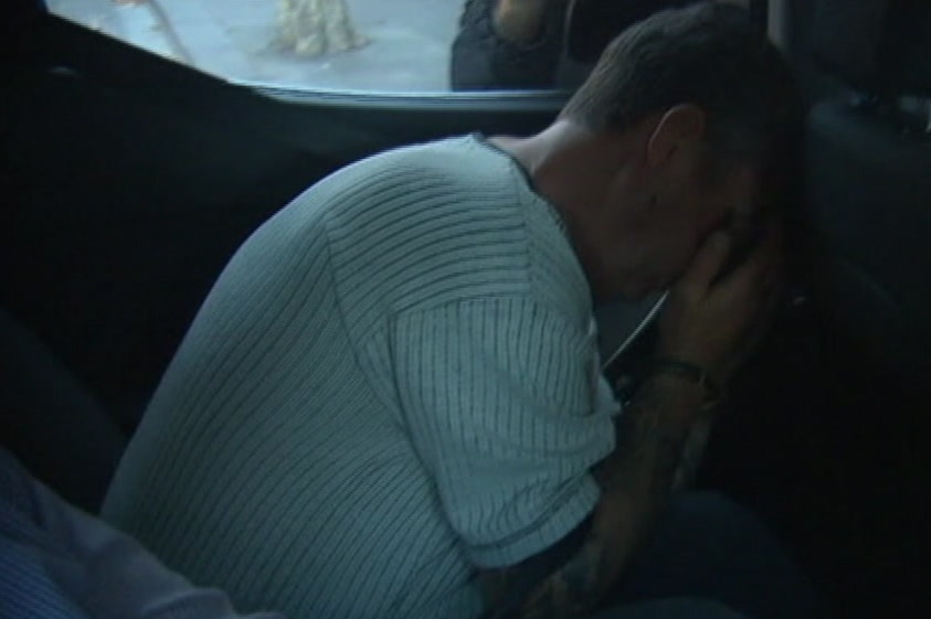 A man seated in a police car concealing his face with his hands.