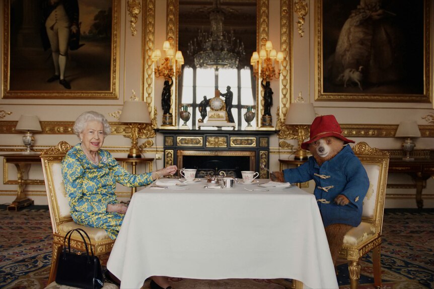 Королева сидит за столом с медвежонком Паддингтоном. 
