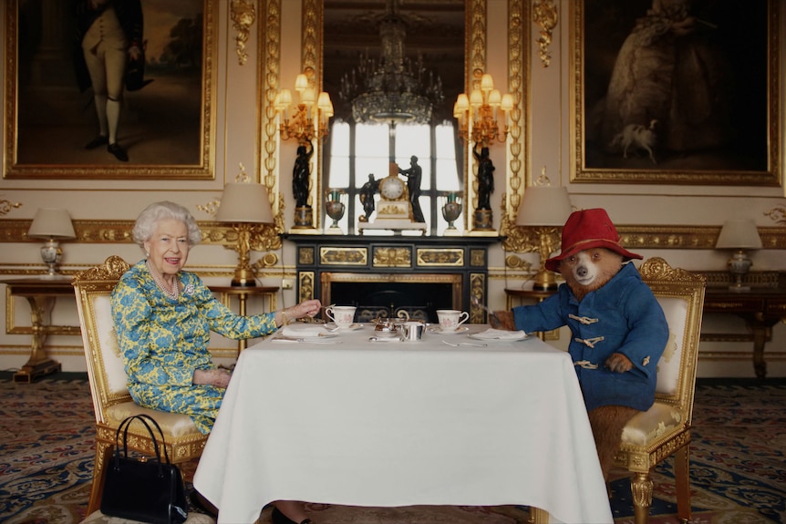 The Queen sitting at a table with Paddington Bear. 