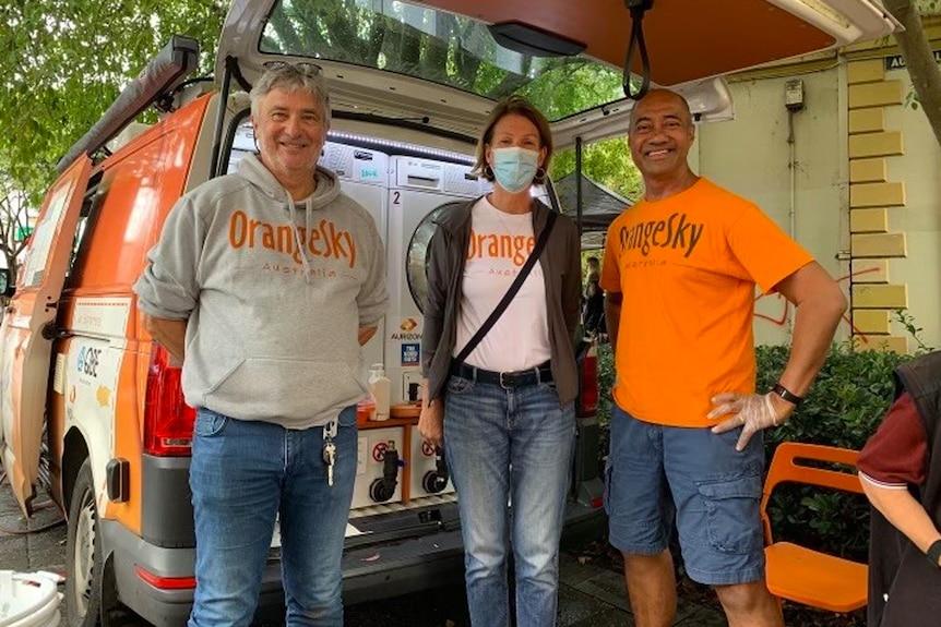 Two men and a woman, in 'Orange Sky' jumpers and t-shirts, pose for a photo in front of a van