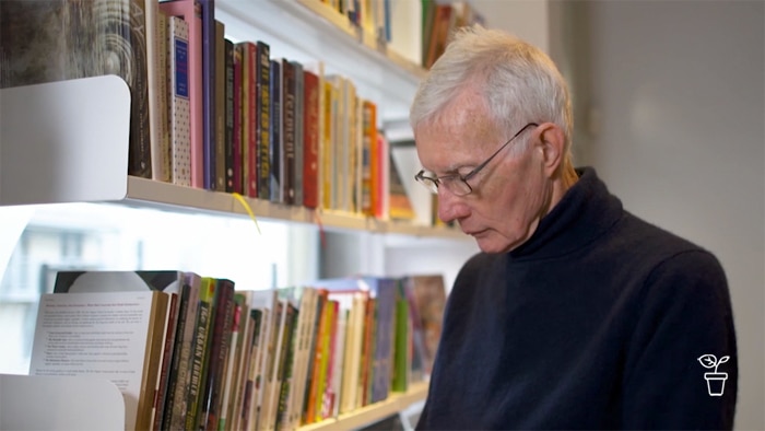 Man with glasses reading a book standing next to filled bookshelf