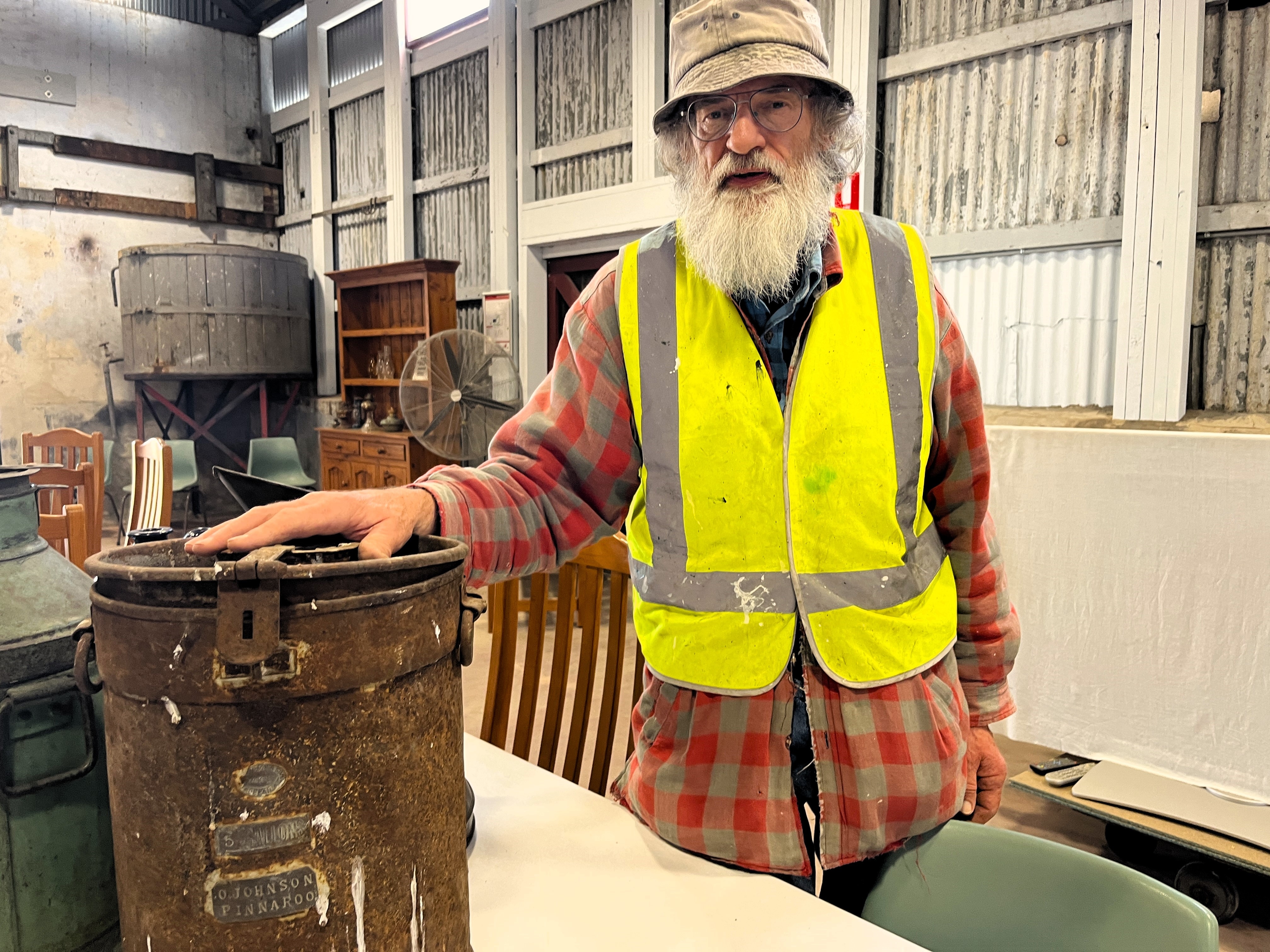 A man with a long beard in a high vis vest rests his hand on top of an aged, rusty milk can with a name tag