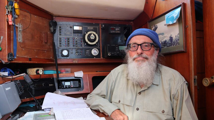 A man sits inside a yacht cabin at a table. Communication gear is on walls around him.