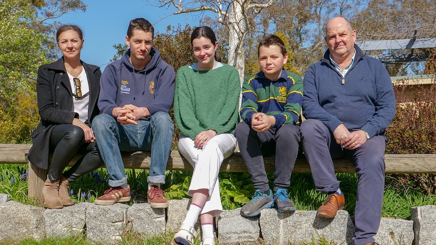 A mum, dad and their three kids - two boys and a teenage girl - sit on a wooden fence.