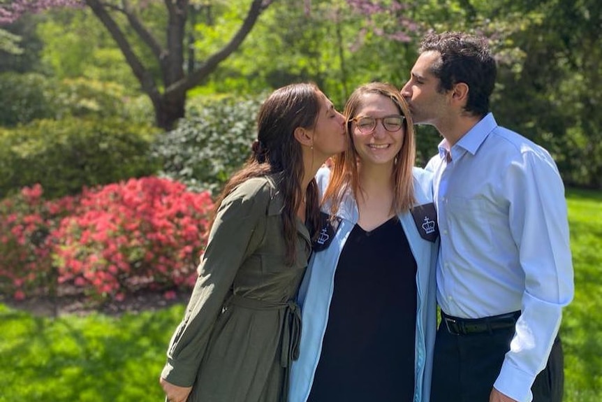 A young woman in glasses smiles as a man and woman proudly kiss her cheeks