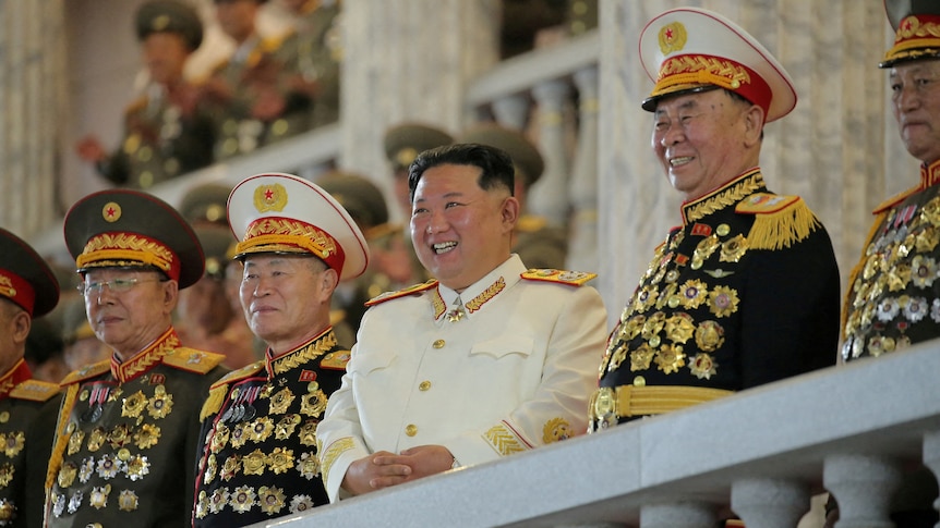 Kim Jong Un smiles as he stands between several army officers on a balcony.