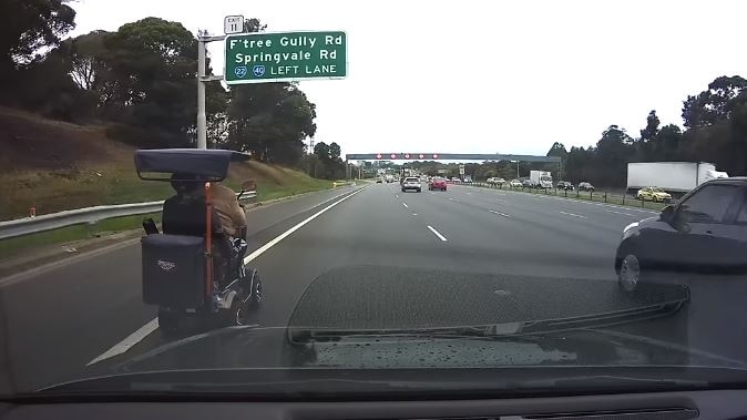 A mobility scooter is driven down a freeway, under a large sign that says 'F'tree Gully Rd, Springvale Rd, Left Lane'.