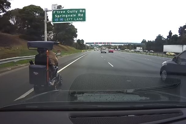 A mobility scooter is driven down a freeway, under a large sign that says 'F'tree Gully Rd, Springvale Rd, Left Lane'.