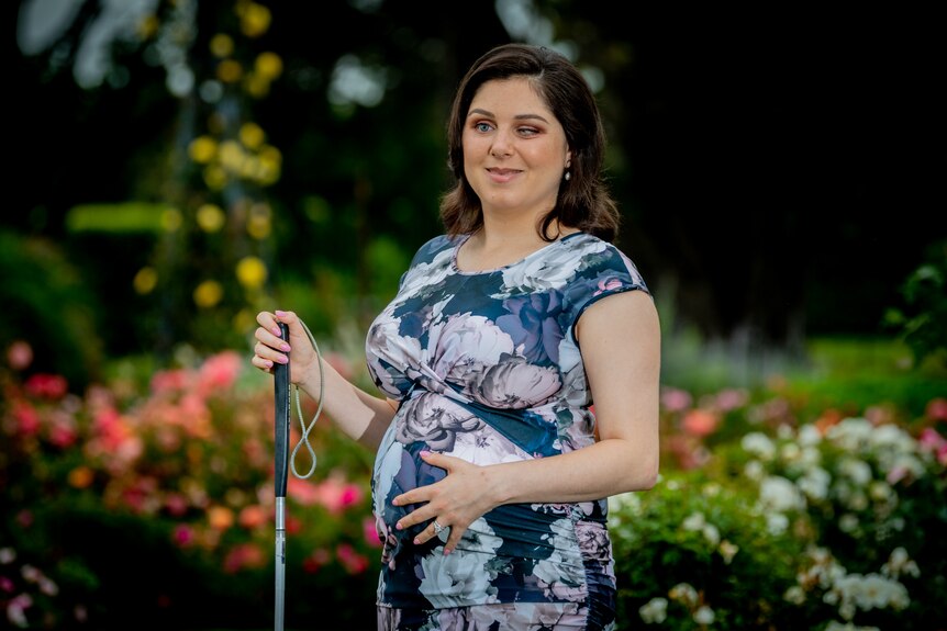 Nas rests one hand on her baby bump and holds a cane with the other, while standing in front of a colourful garden.