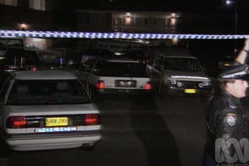 Police cars line the streets as an officer raises police tape to let a car through.