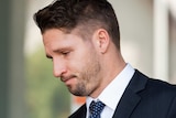 A tight side-on head and shoulders shot of Jesse Hogan frowning wearing a suit and tie.