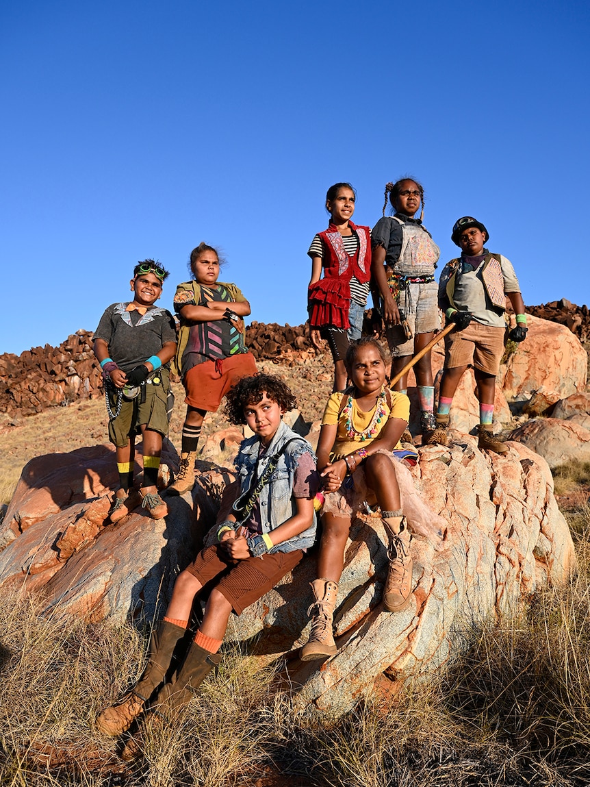 Six tweens stand and sit on large rocks looking into the distance in rocky spinifex desert landscape on sunny clear blue sky day