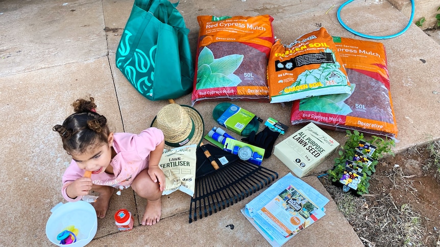 A little girl squats on the ground next to several garden items