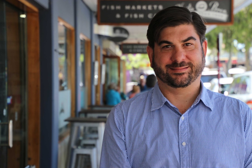 A man with dark parted hair and a beard and blue checked shirt stands on a cafe strip